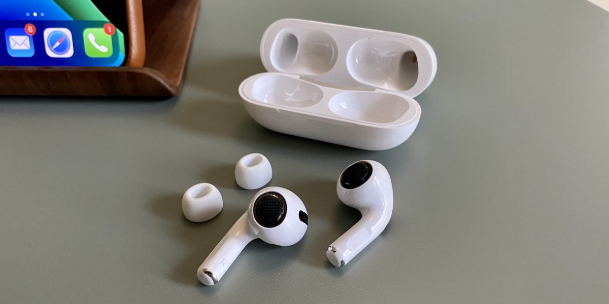 Sepasang AirPods Pro, dengan tips yang terlepas dari AirPods, di belakangnya terdapat tempat AirPods Pro.